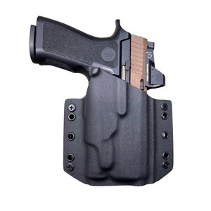 May include: Black handgun with a tan slide and a red dot sight, secured in a black holster. The holster has multiple mounting holes and is designed for secure carry. The firearm is set against a white background.