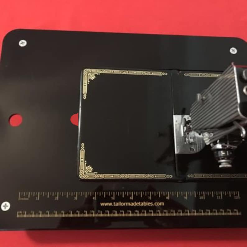 May include: Black sewing machine with a gold trim on a black surface with a ruler that reads "www.tailormadetables.com".