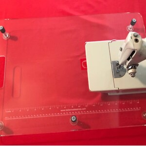 May include: A clear acrylic sewing table with a ruler and the text "www.tailormadetables.com". A white sewing machine is on the table.
