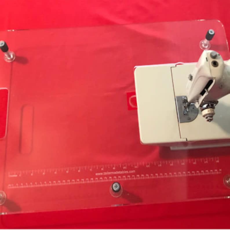 May include: A clear acrylic sewing table with a ruler and the text "www.tailormadetables.com". A white sewing machine is on the table.