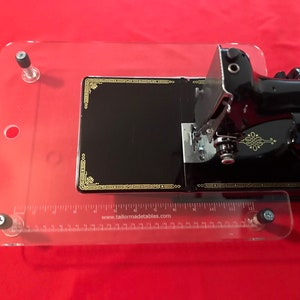 May include: A clear acrylic sewing machine table with a ruler and four screws. The table is designed to hold a small sewing machine. The ruler has markings in inches and the text "www.tailormadetables.com".