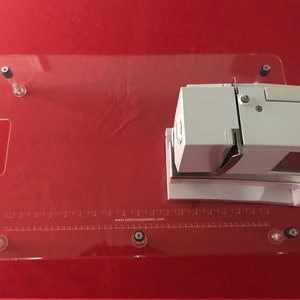 May include: A clear acrylic sewing table with a ruler and the website address "www.tailormadetables.com" printed on it. The table has a handle and is attached to a white sewing machine.