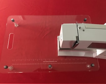 May include: A clear acrylic sewing table with a ruler and the website address "www.tailormadetables.com" printed on it. The table has a handle and is attached to a white sewing machine.