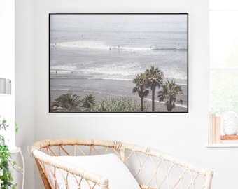 May include: A framed photograph of a beach scene with palm trees and surfers in the ocean. The photo is hanging on a white wall above a wicker chair.