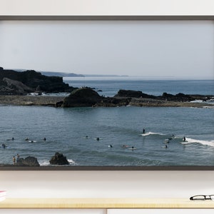 Può includere: Una vista panoramica di una costa rocciosa con surfisti in acqua. L'acqua è di un blu chiaro e il cielo è di un grigio chiaro. Le rocce sono di un marrone scuro.