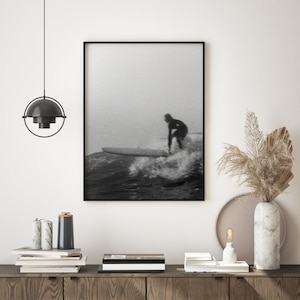 K&ouml;nnte beinhalten: Schwarzwei&szlig;foto eines Surfers, der auf einer Welle reitet. Das Foto ist in einem schwarzen Rahmen gerahmt und h&auml;ngt an einer wei&szlig;en Wand.