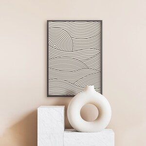Pu&ograve; includere: Una stampa incorniciata nera di un design geometrico di arte lineare con onde astratte in color crema. Un vaso in ceramica bianca con un taglio circolare si trova su un piedistallo bianco.