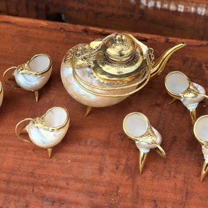 May include: A set of six gold and white shell teacups and teapot. The teapot has a gold lid and a gold handle. The teacups have three legs each.