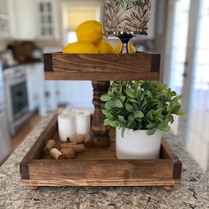 May include: A two-tiered wooden tray with a distressed brown finish. The top tier features a silver candle holder with a leafy design and lemons. The bottom tier has white candles, wine corks, and a potted green plant.