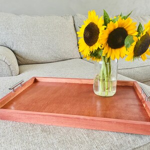 May include: A rectangular wooden serving tray with a reddish-brown finish and silver-toned handles. The tray holds a clear glass jar with sunflowers. The sunflowers have bright yellow petals and dark brown centers. The tray is on a light-colored upholstered ottoman.