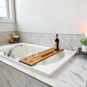 May include: A white bathtub with a wooden bath tray, a glass, and a bottle of wine. The bathtub is in a bathroom with white marble tile.