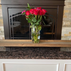 May include: A wooden tray with turned legs sits on a granite countertop in front of a fireplace. A glass vase with red roses sits on the tray.