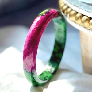 May include: A colourful gemstone bangle bracelet. The bracelet transitions from a deep magenta to a speckled green hue. The smooth, rounded design of the bracelet is displayed against a soft, white background.