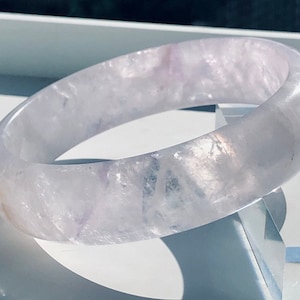 Puede incluir: Un brazalete de amatista lavanda pálido con un acabado liso y pulido. El brazalete está hecho de amatista natural y tiene un aspecto delicado y femenino.