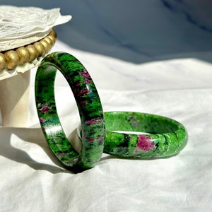 May include: Two green and pink gemstone bangles. The bangles are made of a polished stone with a swirling pattern of green, pink, and black.