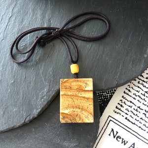 May include: A square-shaped pendant made of brown and white stone with a brown cord necklace. The pendant has a light brown color with white streaks and a yellow bead at the top.