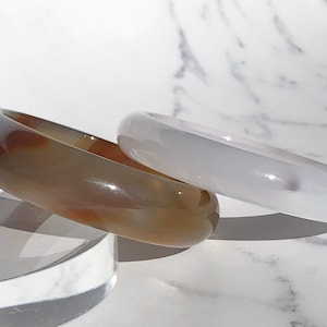 Puede incluir: Dos pulseras de ágata, una marrón con remolinos blancos y naranjas y otra blanca con un toque de morado. Las pulseras están apiladas una encima de la otra sobre una superficie de mármol blanco.