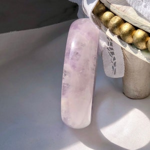Puede incluir: Un brazalete de amatista lavanda pulido y liso. El brazalete es de color morado claro con vetas blancas.
