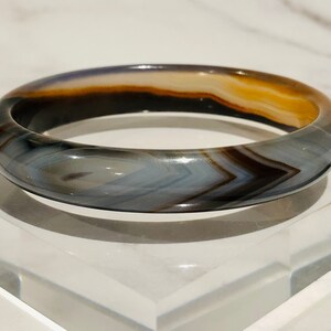 May include: A grey, brown and white agate bangle bracelet. The bracelet is made of polished stone and has a smooth, rounded surface.