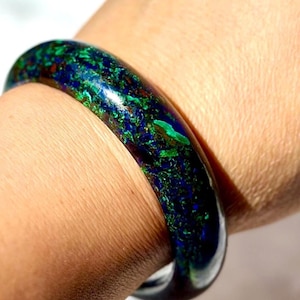 Puede incluir: Primer plano de una pulsera redonda y pulida. La pulsera es una mezcla de azules profundos, verdes y toques de rojo, creando un efecto veteado. La joya se lleva en la muñeca, mostrando su superficie lisa y brillante.