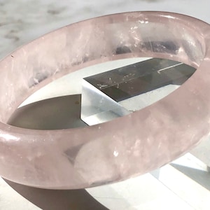 May include: A pale pink rose quartz bangle bracelet. The bracelet is smooth and polished, with a translucent appearance.