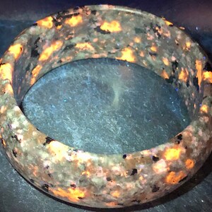 May include: A round, gray and black stone bracelet with orange and yellow flecks. The bracelet is made of a natural stone and has a smooth, polished finish.