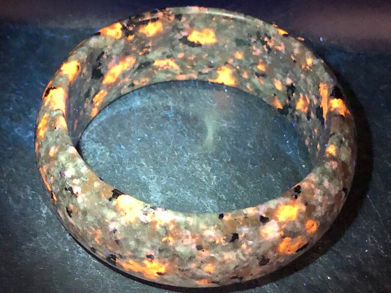 May include: A round, gray and black stone bracelet with orange and yellow flecks. The bracelet is made of a natural stone and has a smooth, polished finish.