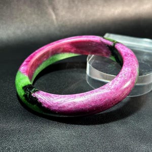 May include: A vibrant bangle bracelet with a polished finish. The bracelet features a striking color combination of pink, green, and black. The gemstone bracelet is a circular shape and appears to be made of a natural stone.