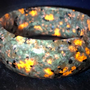 May include: A green and brown speckled bangle bracelet with orange and blue flecks. The bracelet is made of polished stone and has a smooth, rounded surface.