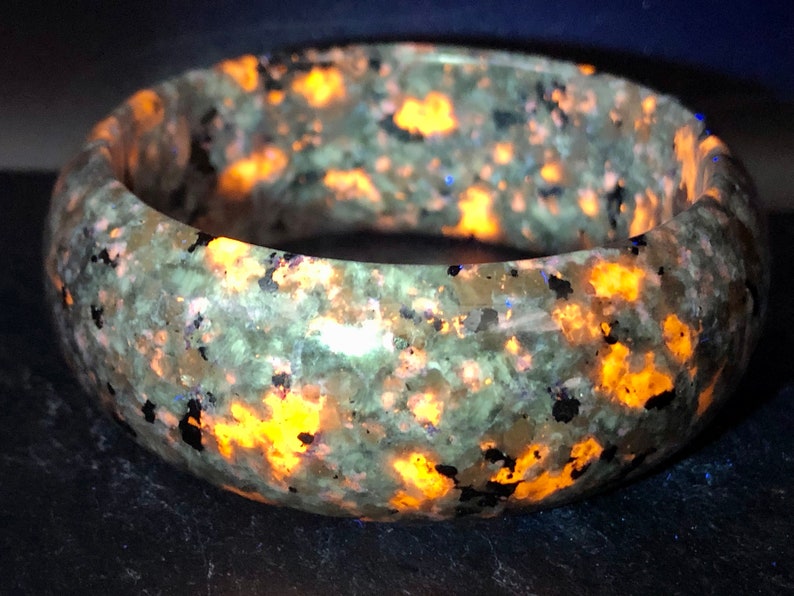 May include: A green and brown speckled bangle bracelet with orange and blue flecks. The bracelet is made of polished stone and has a smooth, rounded surface.