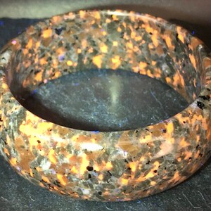 May include: A brown and orange speckled bangle bracelet made of polished stone. The bracelet has a smooth, rounded surface and is approximately 2 inches wide.