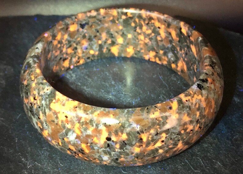 May include: A brown and orange speckled bangle bracelet made of polished stone. The bracelet has a smooth, rounded surface and is approximately 2 inches wide.