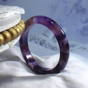 May include: A polished amethyst bangle bracelet. The bracelet is a deep purple color with lighter purple and brown streaks. The smooth, rounded shape of the bracelet is displayed against a white fabric background.