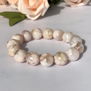 Puede incluir: Un brazalete de ágata blanca con remolinos rosas y marrones. El brazalete está hecho de cuentas redondas y tiene aproximadamente 18 cm de largo.