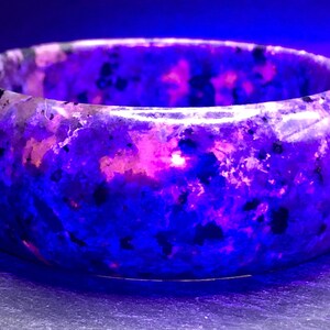 May include: A purple and pink gemstone bangle bracelet with a speckled pattern. The bracelet is made of polished stone and has a smooth, rounded surface.