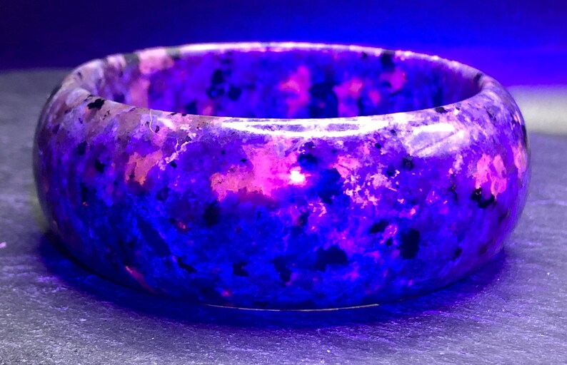 May include: A purple and pink gemstone bangle bracelet with a speckled pattern. The bracelet is made of polished stone and has a smooth, rounded surface.