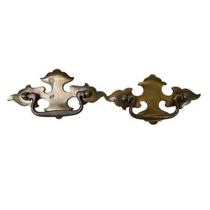 May include: Two antique brass drawer pulls with a decorative cloud-like design. Each pull features a curved handle and is mounted on a decorative plate. The pulls are made of brass and have a vintage aesthetic.