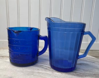 May include: Two vintage cobalt blue measuring cups. The smaller cup has measurements in cups and ounces. The larger pitcher has a handle and a spout. Both are made of a translucent material and sit on a marble-like surface.