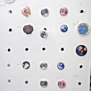 May include: A collection of decorative buttons with various designs, including a swan, a bunny, and floral patterns. The buttons are in various colors, such as blue, pink, and white, and are arranged on a white surface.