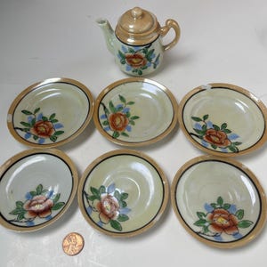 May include: A miniature porcelain tea set consisting of one teapot with a lid and six matching saucers. Each piece features a cream-colored base with a hand-painted orange flower, green leaves, and blue accents, finished with a metallic gold rim.