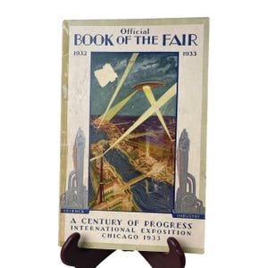 May include: Vintage "Official Book of the Fair" from the 1933 Chicago World's Fair. The cover features an illustration of the fairgrounds with a futuristic design, including a flying saucer and beams of light. The book is held by a dark brown stand.