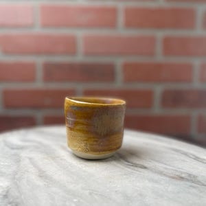 May include: A small, handmade ceramic cup with a textured, glazed surface. The cup features a gradient of colors, transitioning from a light beige base to shades of yellow and brown at the top. The cup is approximately 3 inches tall.