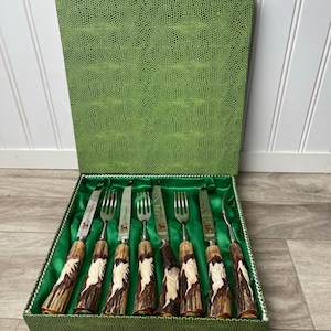 May include: A set of vintage cutlery, including forks and knives, presented in a green textured box with a satin lining. The handles are carved with a brown and cream design. The blades are silver.