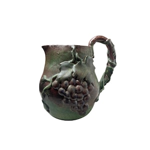 May include: A handcrafted ceramic pitcher with a dark green and brown glaze. The pitcher features a detailed relief of grapes and leaves, with a handle designed to resemble a vine. The pitcher is approximately 7 inches tall.
