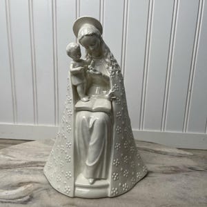 May include: A white ceramic figurine depicting a woman with a halo holding a child. The woman is seated, wearing a long robe with a floral pattern. The child stands on an open book, also holding a flower.