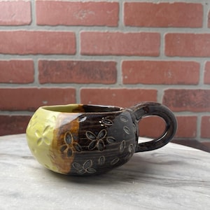 May include: Handmade ceramic mug with a unique design. The mug features a rounded shape with a handle and is divided into three sections: yellow, brown, and dark brown. The dark brown section is decorated with floral patterns. The mug is suitable for hot beverages.