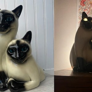 May include: Two Siamese cat figurines. The cats have cream-colored bodies, black faces, ears, and paws, and striking blue eyes. One cat is seated, and the other is standing behind it. The figurines are ceramic and have a glossy finish.