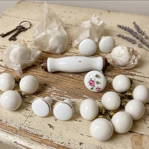 May include: A collection of vintage cabinet knobs and pulls in various styles. The knobs are primarily white ceramic with brass accents, some featuring gold rims or floral designs. A white ceramic pull and antique keys are also present.