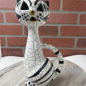 May include: A ceramic cat figurine with a white crackle glaze and black accents. The cat has black stripes, spots, and ear details. The eyes are dark green. The figurine is sitting on a marble surface.