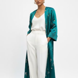 May include: A teal-colored, long, open-front robe with a floral pattern. The robe is worn over a white top and white pants. The model is wearing gold hoop earrings and a gold necklace.